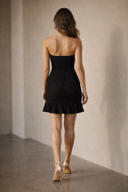 Woman wearing a black strapless heart textured dress with ruffled hem, standing against a neutral wall.