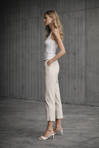 Woman wearing a white tank top and beige capri pants standing against a concrete wall.