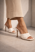 White high-heeled shoes worn with beige pants on a neutral background