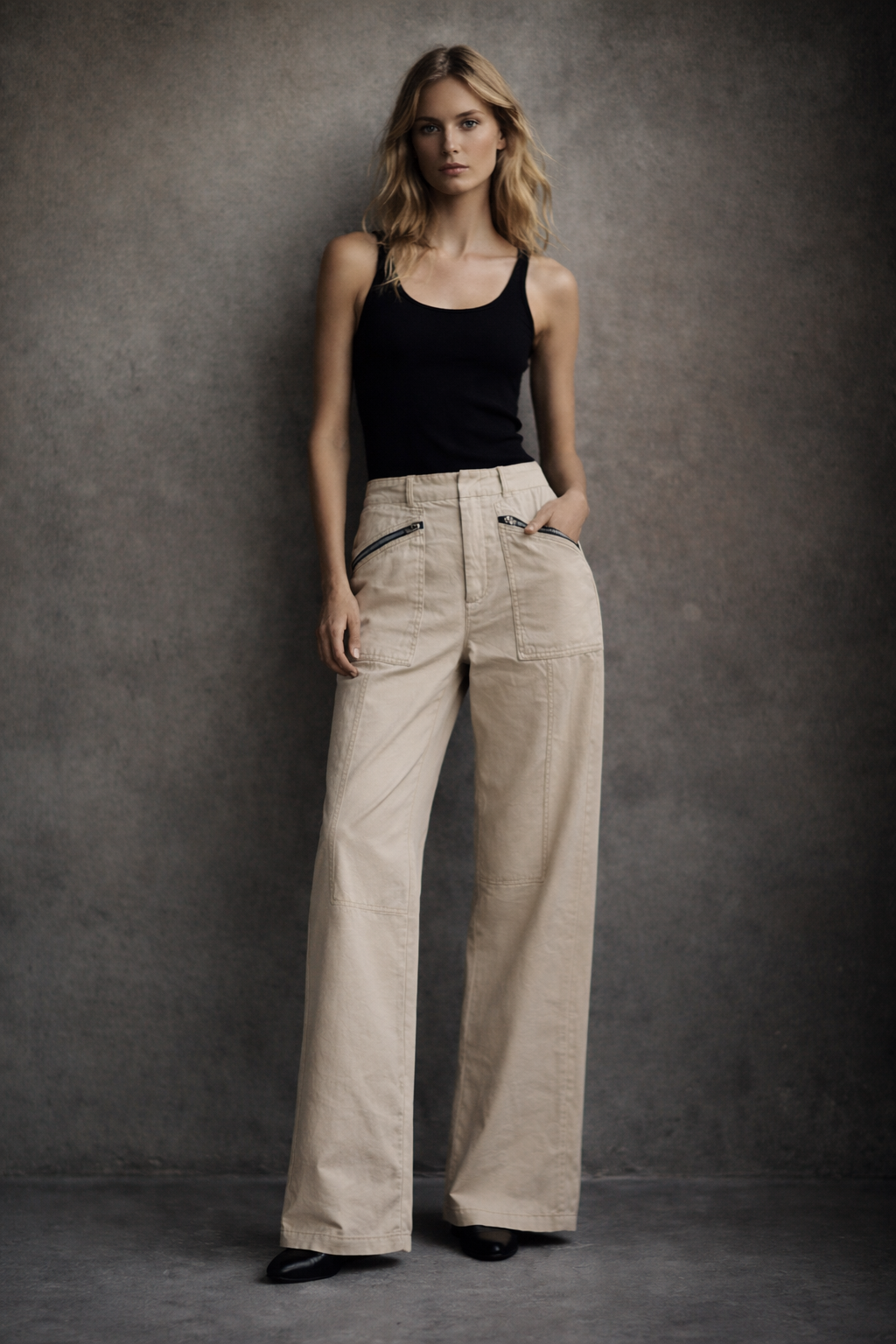 Woman wearing a black tank top and beige pants against a gray background