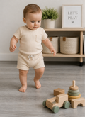 Baby in a beige outfit standing next to wooden toys in a room with a shelf and plants.