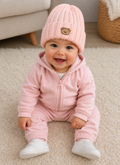 Baby in pink outfit with a matching hat, sitting on a carpeted floor.
