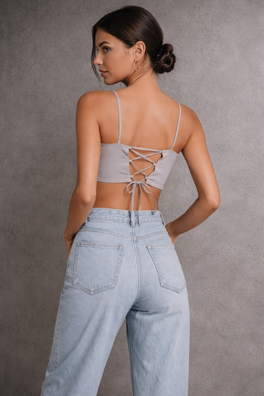 Woman wearing a gray crop top with lace-up back and light blue jeans against a gray background