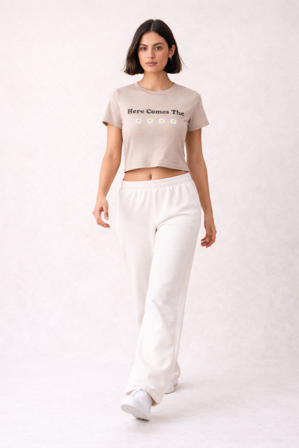 Woman wearing a beige graphic t-shirt here comes the sun and white pants on a white background