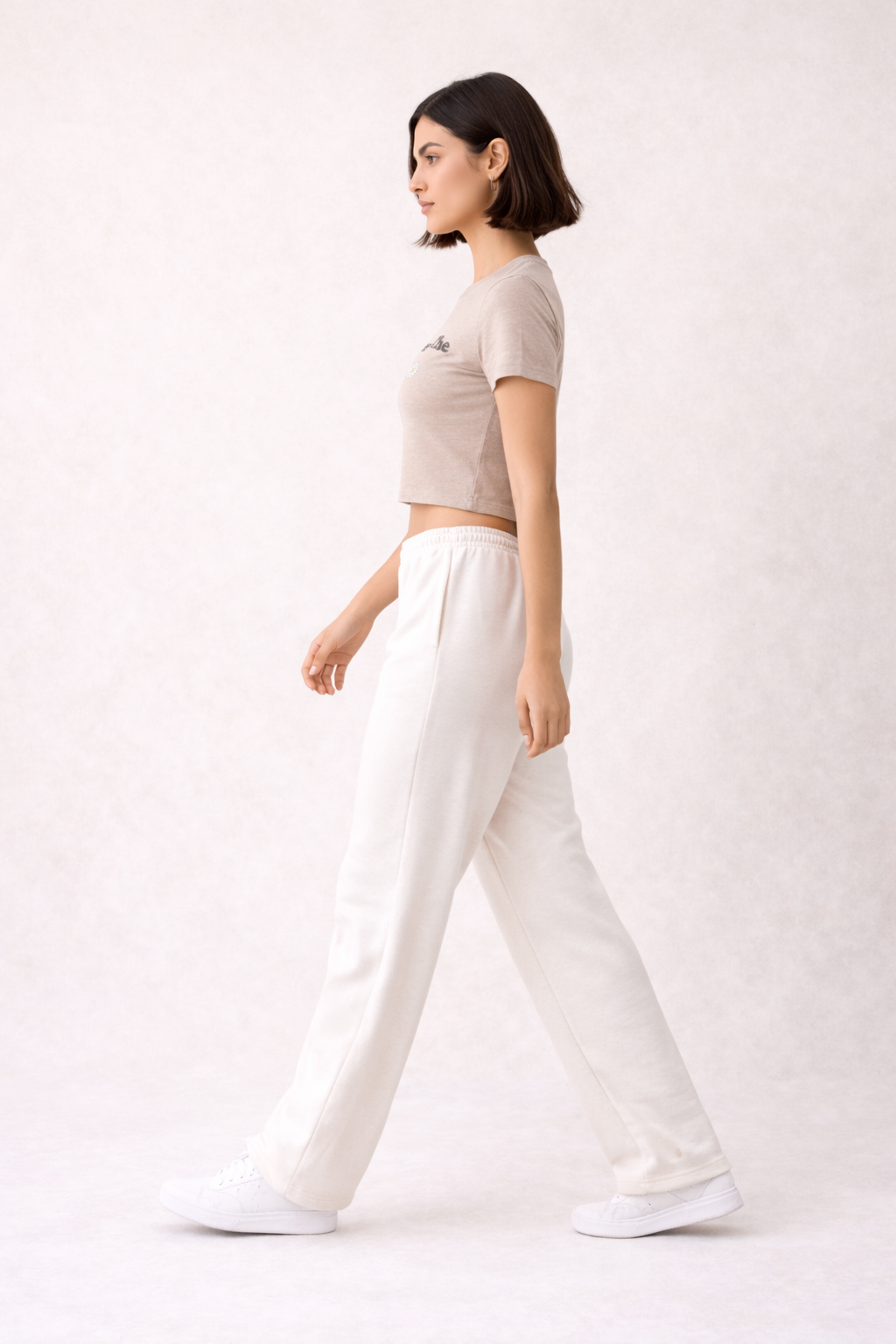 Woman wearing a beige graphic here comes the sun crop top and white pants on a white background