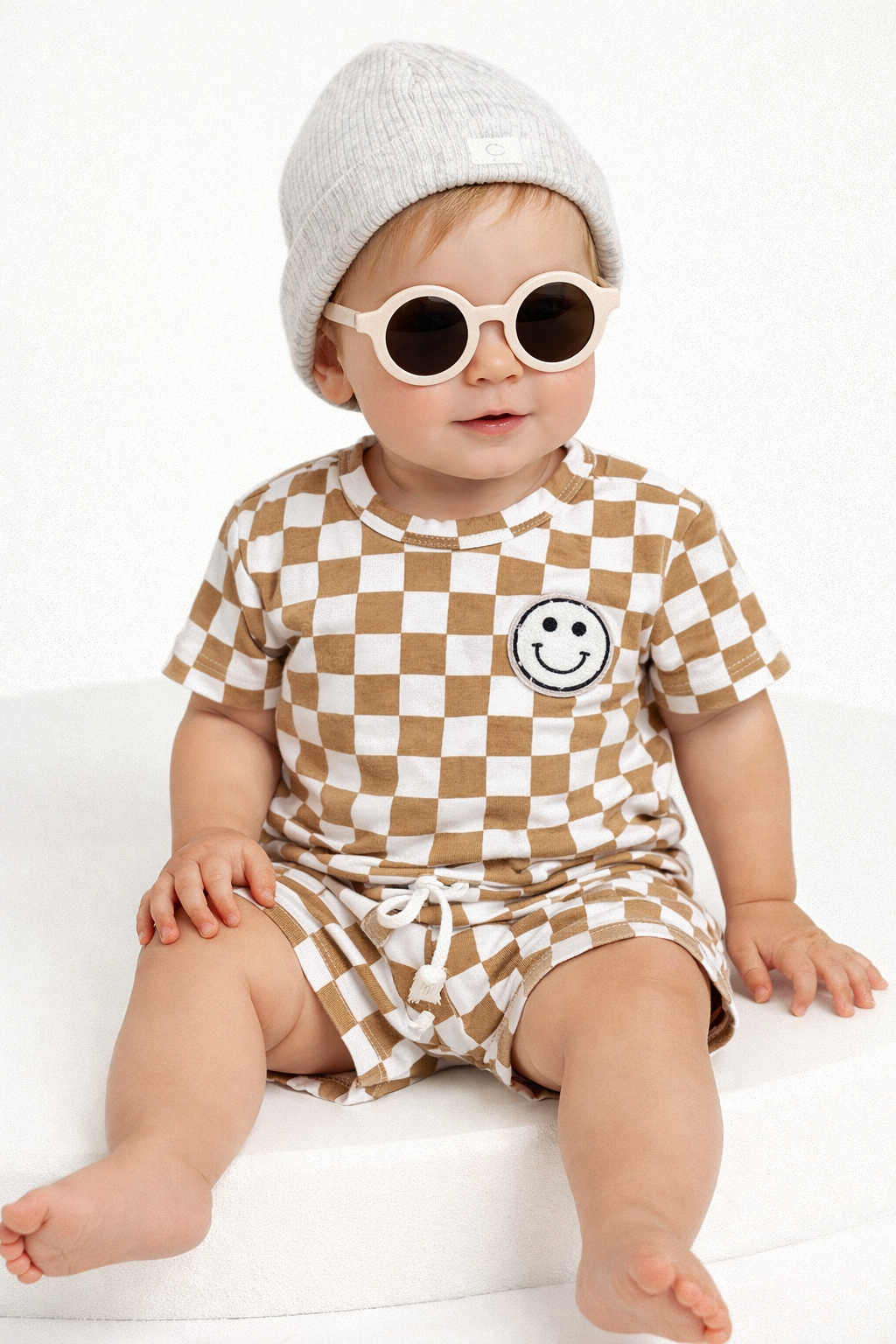 Baby wearing a checkered outfit with a smiley face, white hat, and sunglasses on a white background